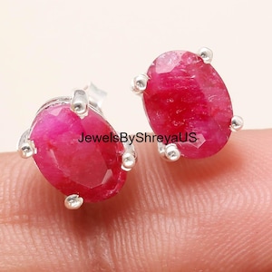 Op de afbeelding: Een paar ovaalvormige robijn oorknopjes gezet in zilveren pootjes. De diep roze-rode edelstenen hebben een gefacetteerd oppervlak. De tekst JewelsByShreyaUS is zichtbaar in de afbeelding. De oorbellen hebben een klassieke vier-pootjes zetting.