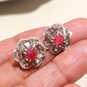 May include: A pair of silver-coloured flower-shaped stud earrings, each with a bright red gemstone centre. The earrings are held in a person's hand. The earrings have a detailed, openwork design around the gemstone.