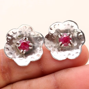 May include: A pair of silver-colored flower-shaped stud earrings. Each earring features a hammered texture and a vibrant red gemstone at the center. The earrings are displayed against a light background.