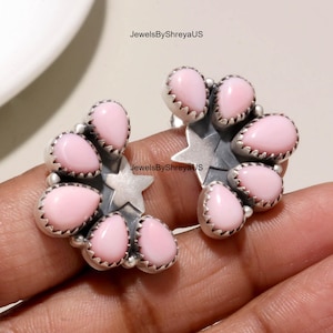 May include: A pair of silver earrings featuring a star-shaped center surrounded by five teardrop-shaped pink stones in serrated bezels. The text JewelsByShreyaUS is visible on the image. The design is symmetrical with a polished metallic finish.