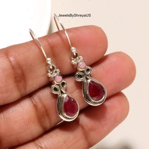 May include: A pair of silver drop earrings featuring a teardrop-shaped dark red gemstone at the base and a small round pink stone above it. The metalwork includes decorative silver loops and hooks. The text JewelsByShreyaUS is visible on the image.