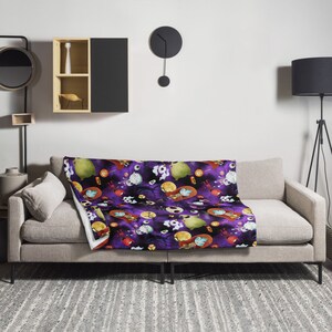 May include: A purple and black throw blanket with a repeating pattern of characters from the Nightmare Before Christmas. The blanket is on a gray couch with a white pillow.