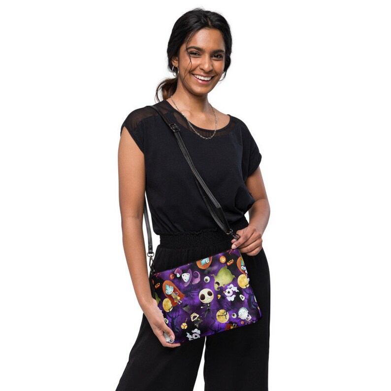 May include: A woman wearing a black top and black trousers is holding a purple and black crossbody bag with a Nightmare Before Christmas print. The bag has a black strap and features characters from the film.