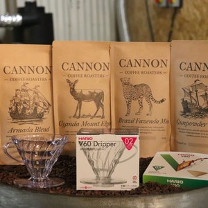 May include: Four Cannon Coffee Roasters coffee bags, including Armada Blend and Gunpowder Blend, with illustrations of a ship, deer, and cheetah. A clear Hario V60 Dripper and filter box are in front, with coffee beans scattered around.
