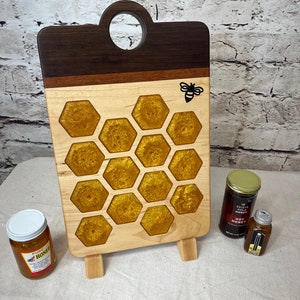 Può includere: Un tagliere in legno con un design a nido d'ape e una silhouette di ape. Il tagliere ha un manico e poggia su tre gambe. È realizzato in legno di noce e acero e ha una riempitura in resina epossidica dorata nel design a nido d'ape.