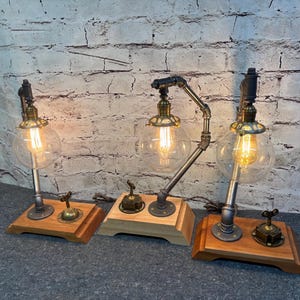 Könnte beinhalten: Drei Tischlampen im Industriestil mit Edison-Glühbirnen. Jede Lampe hat einen transparenten Glaskugel-Schirm, einen Metallsockel und eine Holzplattform. Die Lampen haben eine Vintage-Ästhetik mit freiliegenden Rohren und Messingakzenten.