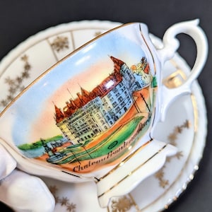 May include: A white porcelain teacup and saucer with a colorful illustration of a castle and the text "Chateau Frontenac" on the side. The saucer has a gold trim and a floral pattern.