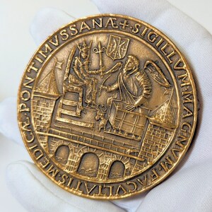 May include: A bronze medal with a raised image of two figures in a castle setting. The inscription around the edge reads "PONTIMVS SANAE + SIGILLVM MAGNVM MEDICVS CALVVS".