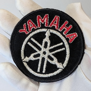 Peut inclure: Un écusson rond noir avec le mot "YAMAHA" brodé en rouge en arc de cercle en haut. En dessous, un emblème blanc. L'écusson est probablement destiné à une moto ou à un autre véhicule.