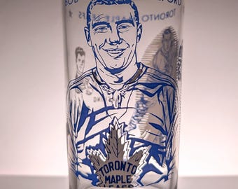 May include: A clear glass tumbler featuring a blue graphic illustration of hockey player Bob Pulford. The design includes the text Bob Pulford and the Toronto Maple Leafs logo with a maple leaf icon. The glass has a cylindrical shape and smooth rim.