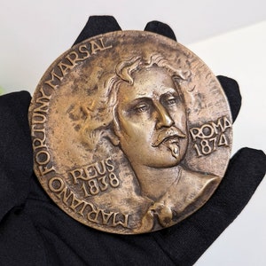 May include: A bronze medal with a portrait of a man with a mustache and beard. The medal is inscribed with the words "MARIANO FORTUNY MARSAL REUS 1838 ROMA 1874".