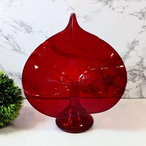 Puede incluir: Un florero de vidrio rojo vibrante con un cuerpo ancho y plano en forma de corazón que se estrecha hacia una punta superior. El recipiente se apoya sobre una base circular y cuenta con una abertura central, con un acabado liso y translúcido.