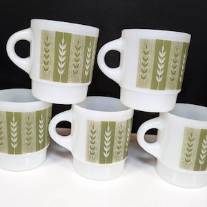 Peut inclure: Un ensemble de cinq tasses en verre opalin blanc, chacune ornée d'un motif vertical répétitif de tiges de blé vert olive. Les tasses présentent une forme cylindrique avec une base étagée et une anse incurvée sur le côté, disposées en groupe.