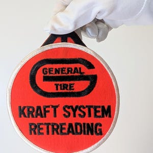 Peut inclure: Un écusson rond rouge avec les mots "GENERAL TIRE" et "KRAFT SYSTEM RETREADING" en noir. L'écusson a une bordure noire et un bord cousu blanc. Une main gantée de blanc tient l'écusson.