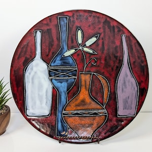 May include: A circular decorative plate featuring a red background and stylized bottle and vase designs in white, blue, orange, and purple. The plate is displayed on a metal stand.