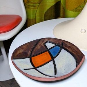 May include: A round ceramic dish featuring a hand-painted abstract design with a dark brown border, a central orange rectangular shape, a blue triangular accent, and off-white sections, all separated by thick, raised black lines.