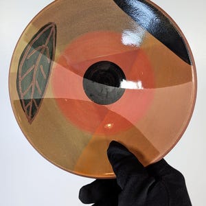 May include: A round ceramic bowl featuring a glossy finish with a central black circle surrounded by concentric bands of orange, tan, and brown. A stylized dark green leaf outline is painted on the left side, and a black arc accents the top edge.