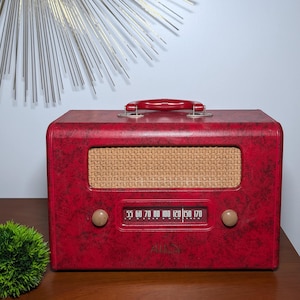 May include: A retro red radio with a woven speaker cover and a red handle. The radio features a dial with numbers and a brand name. The radio is on a wooden surface.