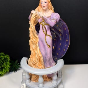 Peut inclure: Figurine en porcelaine d'une femme aux longs cheveux blonds, vêtue d'une robe lavande avec des détails dorés et d'une cape bleu foncé avec des étoiles dorées. La figurine est posée sur une base blanche en forme de balcon.