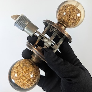 Peut inclure: Un ensemble de poignées de porte composé de deux boutons sphériques en verre transparent remplis de flocons métalliques dorés. Le mécanisme comprend une tige centrale en métal, deux plaques circulaires couleur bronze et un loquet argenté.