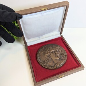 May include: A bronze medal in a brown velvet box with a red velvet lining. The medal features a portrait of a man with the text "TADIANO VICTORIUS MARSAL REUS 1838 ROMA 1874" around the edge.