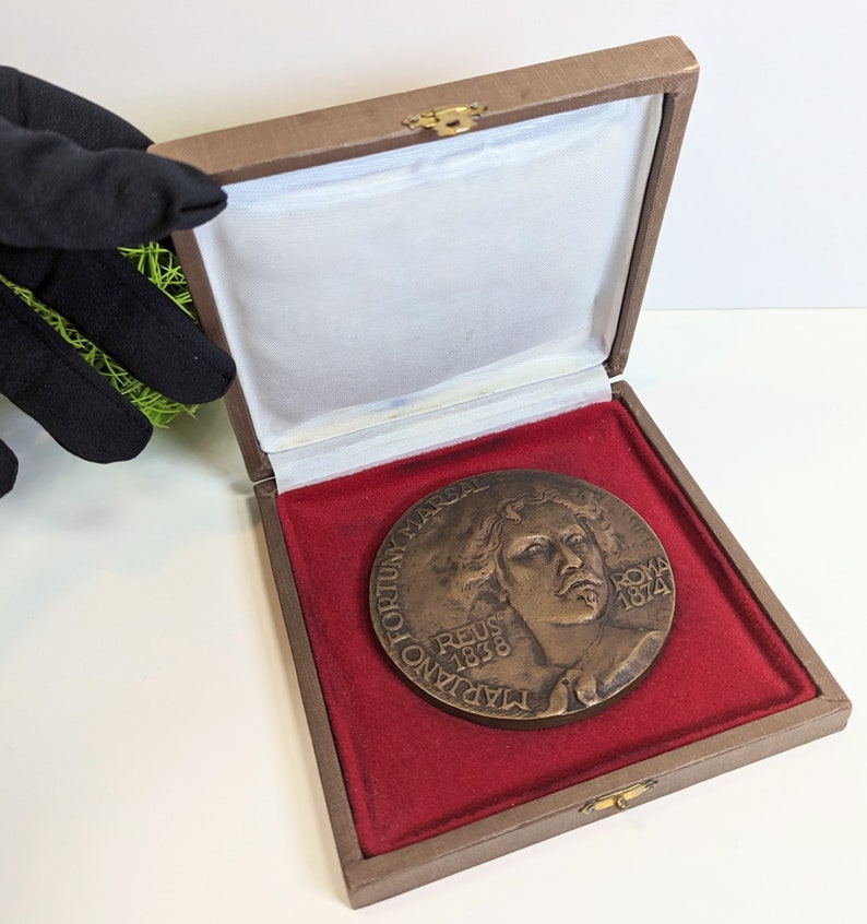 May include: A bronze medal in a brown velvet box with a red velvet lining. The medal features a portrait of a man with the text "TADIANO VICTORIUS MARSAL REUS 1838 ROMA 1874" around the edge.