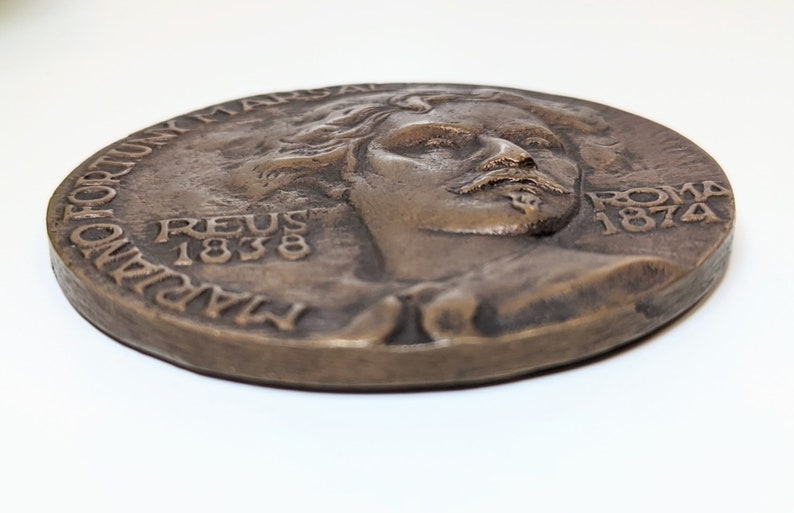May include: A bronze medal with a portrait of a man with a beard and mustache. The medal is inscribed with the text "MARIANO FORTUNY MARSAL REUS 1838 ROMA 1874".