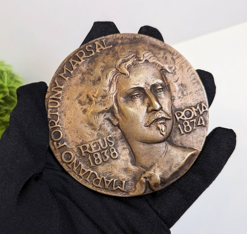 May include: A bronze medal with a portrait of a man with a mustache and beard. The medal is inscribed with the words "MARIANO FORTUNY MARSAL REUS 1838 ROMA 1874".