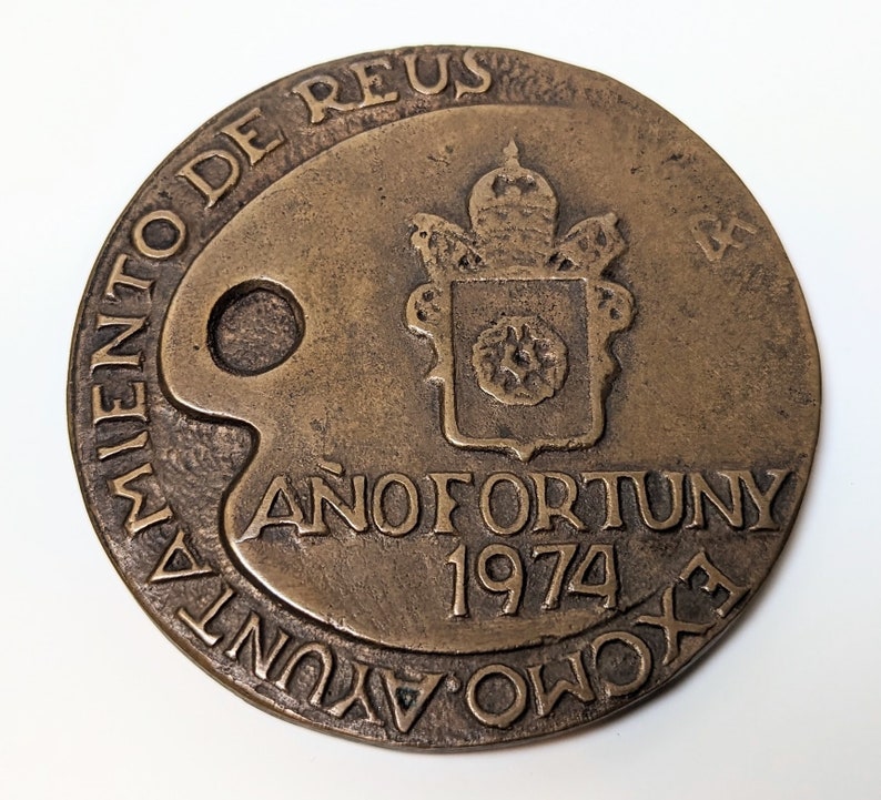 May include: A bronze medal with a papal coat of arms and the text "A&ntilde;o Fortuny 1974" and "Movimiento de Reus" around the edge.