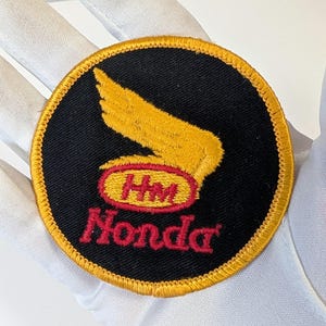 Peut inclure: Écusson rond noir avec un motif d'aile jaune et le texte "HM Honda" en rouge. L'écusson a une bordure jaune et est probablement un emblème ou un logo.