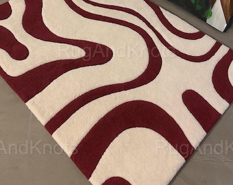 May include: A rectangular rug with a cream-colored background and a bold, wavy pattern in a deep burgundy color. The rug's design features flowing, organic shapes that create a visually dynamic surface. The texture appears soft and plush.