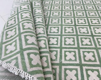 May include: A rolled-up green and white geometric patterned rug with a fringe edge. The rug features a repeating pattern of white crosses within green squares. The design is visible on the rolled portion and the unfolded section.