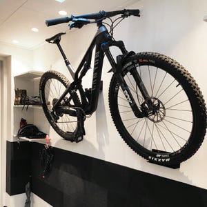Pode incluir: Uma bicicleta de montanha preta com um quadro branco está montada numa parede. A bicicleta tem um selim preto, um guiador preto e rodas pretas com pneus Maxxis. A bicicleta está pendurada num suporte de parede.