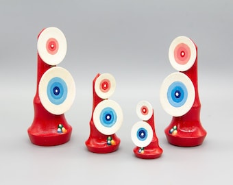 May include: Four red wooden sculptures with white, blue, and pink concentric circles. The sculptures are stacked on top of each other and are of varying sizes.