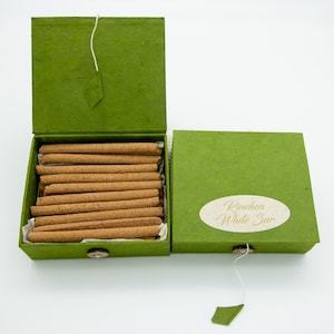 Op de afbeelding: Een groene doos met een open deksel, waarin een stapel bruine wierookstokjes te zien is. De doos heeft een label met de tekst "Rinchen White Sur".