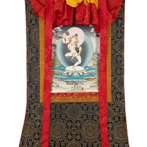 Könnte beinhalten: Ein traditionelles tibetisches Thangka-Gemälde, das eine Gottheit in einem farbenfrohen, kunstvollen Design darstellt. Das Gemälde ist mit einem roten, gelben und goldenen Rahmen versehen und auf einem dunkelblauen Stoff mit goldenem Muster montiert.
