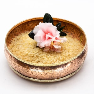 Könnte beinhalten: Eine Kupferschale mit einem verzierten Design, gefüllt mit ungekochtem weißem Reis und mit einer einzelnen rosa Blume verziert.