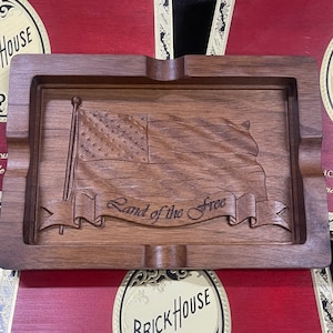 May include: A wooden cigar ashtray with an American flag design and the words "Land of the Free" carved into it. The ashtray is sitting on top of a red cigar box with gold lettering that reads "Brick House - J.C. Newman Cigar Co. - Handmade in Nicaragua".