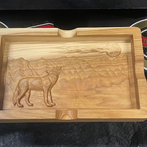 May include: A wooden tray with a carved wolf howling at the moon in a mountain landscape. The tray is made of light brown wood and has a natural finish.
