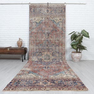 Puede incluir: Una alfombra rectangular grande con un patrón desgastado en tonos rojo terracota, azul apagado y crema. La alfombra muestra un diseño de medallón central rodeado por intrincados bordes florales y geométricos que cubre el suelo.