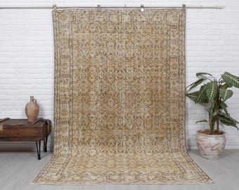 May include: A large, rectangular, vintage rug with an intricate, all-over floral pattern in shades of beige and brown. The rug is displayed against a white brick wall, showcasing its aged appearance and detailed design. The rug's edges are neatly finished.