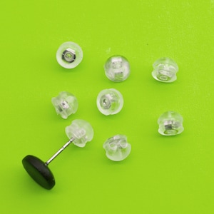 May include: A collection of clear, plastic earring backs and a single earring post with a black backing, set against a bright green background. The earring backs are small, rounded, and translucent, designed to secure earrings.
