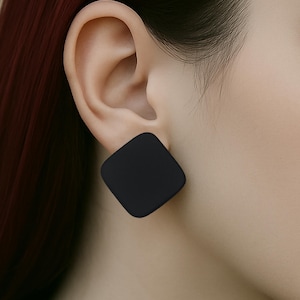 May include: A close-up of a black square earring. The earring is a solid, matte black color and has rounded corners. The earring is worn on the ear of a person with fair skin and dark red hair.