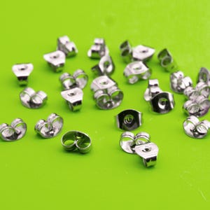 May include: A selection of silver-coloured earring backs, displayed on a vibrant green background. The earring backs are in various shapes, including butterfly and post styles, and are made of a shiny metal.