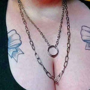 May include: Two silver chain necklaces with a large silver ring pendant. The necklaces are layered on a person's chest. The person has two tattoos on their chest, one of a blue bow and the other of a pink bow.