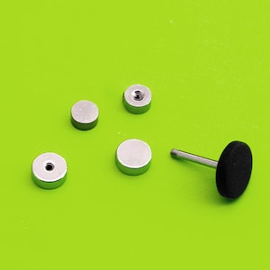 May include: Several silver-coloured, round ear studs and a single black, round ear stud with a silver post. The studs are arranged on a bright green background. The ear studs are small and simple in design.