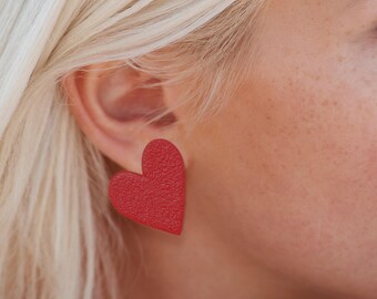 May include: A close-up of a red heart-shaped earring. The earring has a textured surface and is worn on the ear of a person with blonde hair. The earring is a bold, eye-catching accessory.