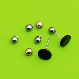 May include: A selection of stud earrings and earring backs against a vivid green backdrop. The earrings feature silver and gold-toned ball studs, and black disc studs. The earring backs are also visible.
