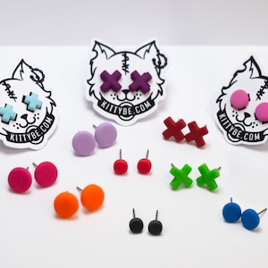 Peut inclure: Une collection de boucles d'oreilles clous colorées de différentes formes, dont des modèles ronds et en croix. Les boucles d'oreilles sont dans les tons de rose, orange, rouge, vert, bleu, violet et noir. L'image présente également des autocollants avec une tête de chat et le texte "KITTYBE.COM".