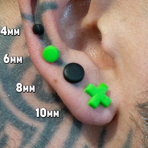 May include: Four different sizes of ear gauges, 4mm, 6mm, 8mm, and 10mm. The gauges are made of black and green plastic. The gauges are shown in a person's ear.